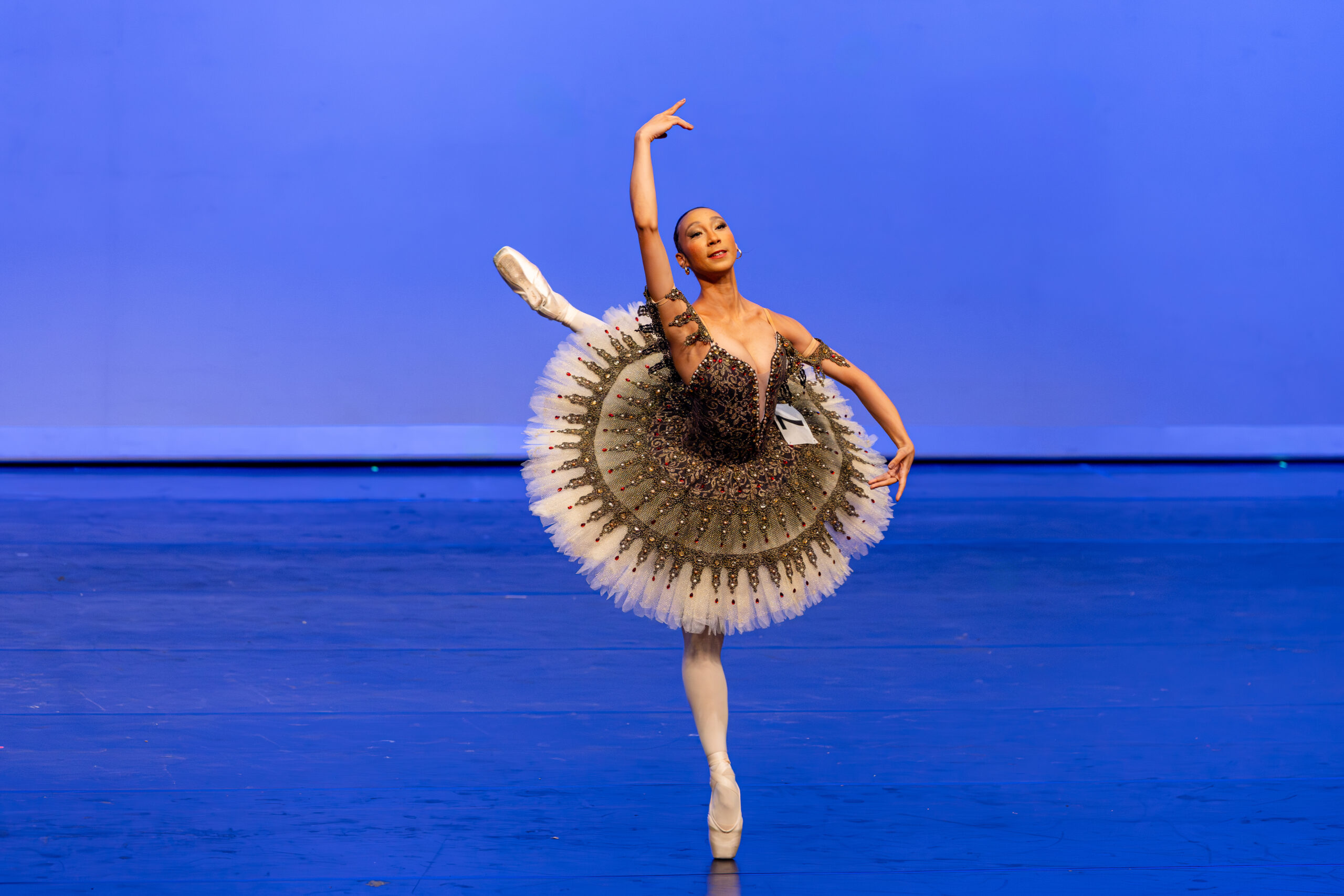 Onstage, Yasmin Oliveira Lopes dos Santos poses on pointe with her back leg in a high attitude derriere. Her arms are in a high fourth position, and she wears a bronze tutu with red and gold detailing. A number is pinned onto her tutu.