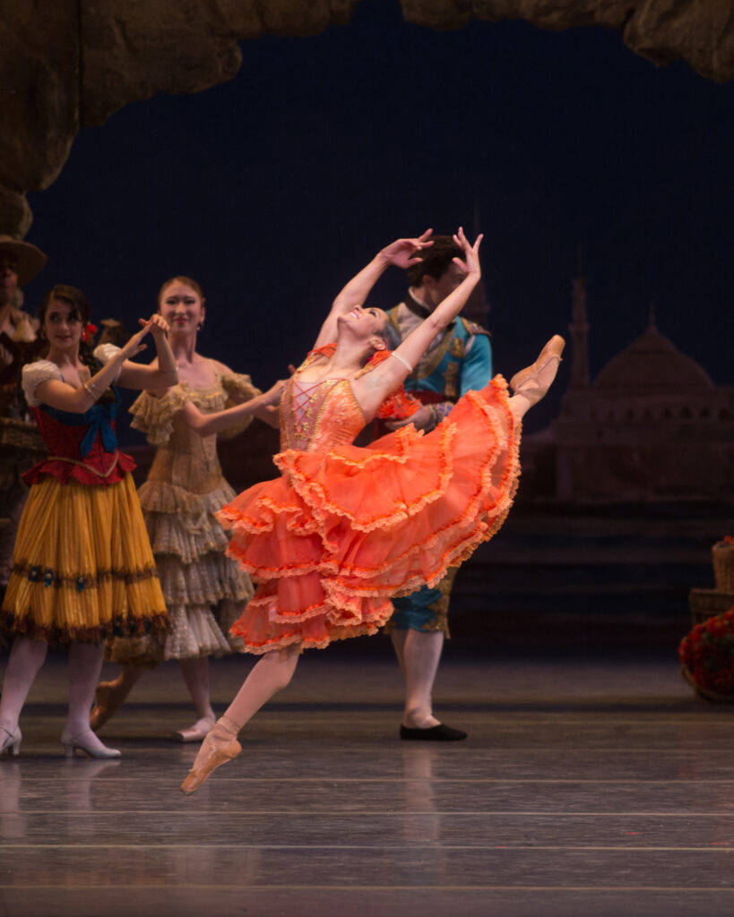 Misty Copeland is captured mid-air in the iconic leap from Don Quixote; she arches her back with her arms in high fifth toward her back leg. She wears a ruffled coral dress with over tights and pointe shoes.