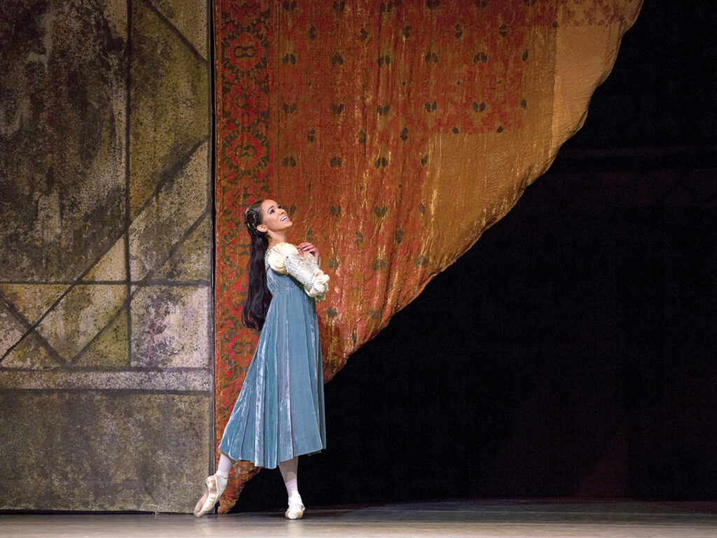 Misty Copeland smiles brightly as she gazes up into the middle distance. She is seen in profile, posed in tendu derrière, hands clasped at her heart. She is costumed in a long, blue velvet gown with puffy white sleeves and pointe shoes; she seems small and young at the foot of a large tapestry set piece.