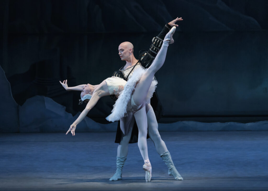 Sara Mearns wears a white tutu and white swan headpiece while developing her right leg to the right while extended back over Tyler Angle's right arm with her arms above her head in a V. Tyler looks down at her with his left arm extended straight in a diagonal to the upper right.
