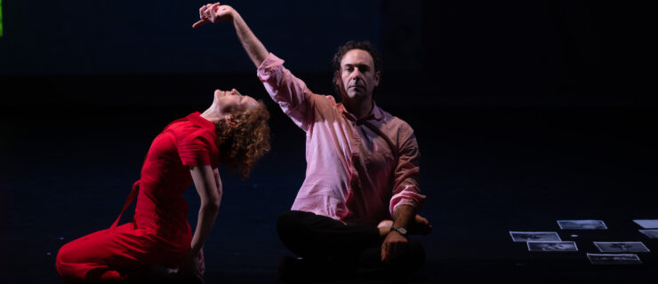 Emily Coates, in red, is shown in profile as she sits on her heels and arches back to face the ceiling. Derek Lucci is seated beside her, looking out at the audience as he raises a lazy hand over her head. Pages with printed photos, not quite visible, are arrayed in a line in front of them.