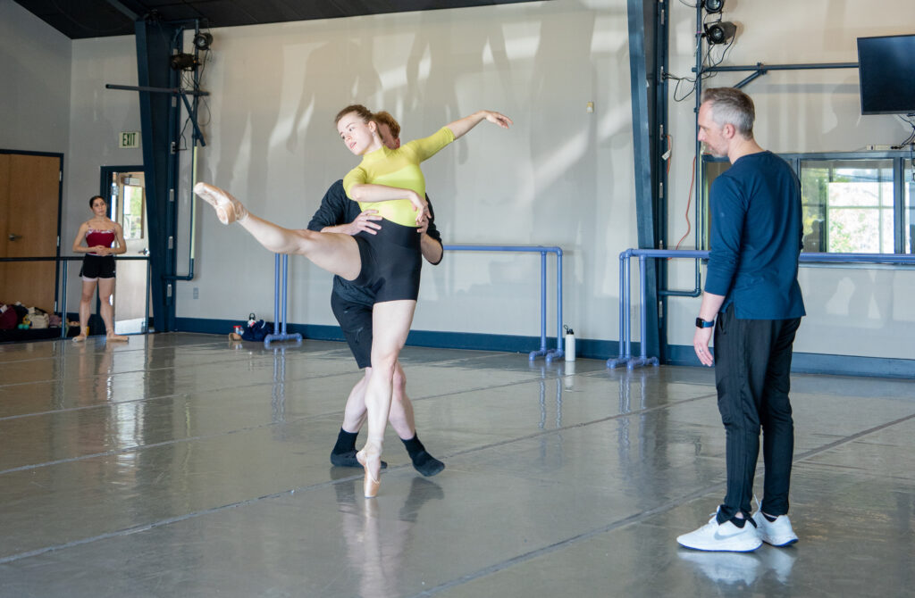 During rehearsal, a dancer en pointe in a yellow leotard and black shorts partners with a male dancer while looking to the left with her left leg in a high second position. Across from her on the right, a rehearsal director wearing white nikes, black joggers, and a navy blue long sleeve shirt watches her.