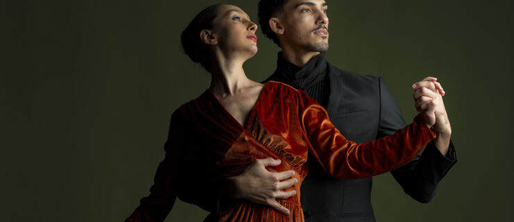 A male dancer looks to the right in a black suit while holding a female dancer in a red velvet dress against his chest with their hands clasped in front of them against a green backdrop.