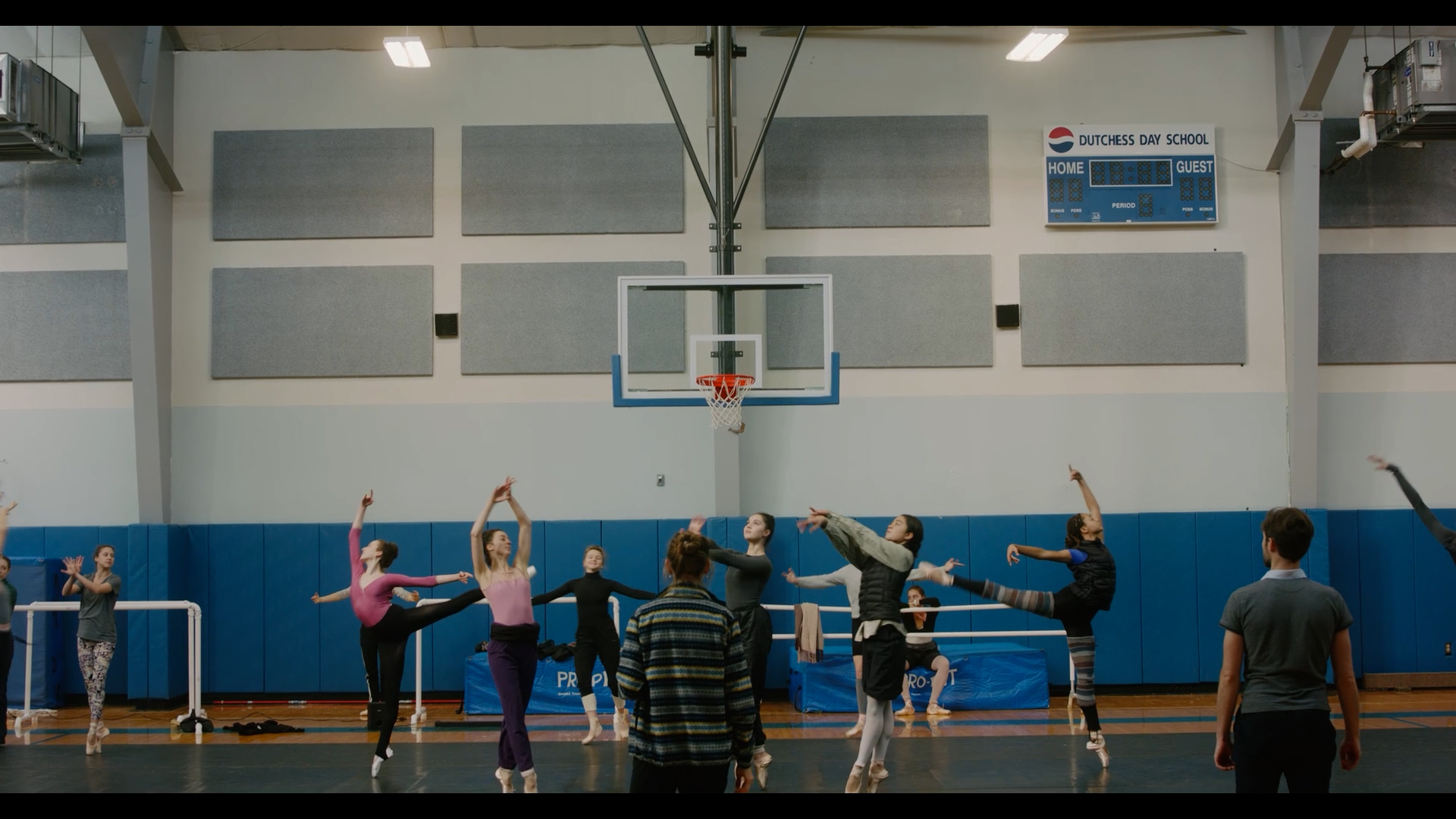 In a school gymnasium, a group of female dancers in practice clothes rehearse a ballet, making various poses. A man and woman, their backs to the camera, stand in front of them and watch.