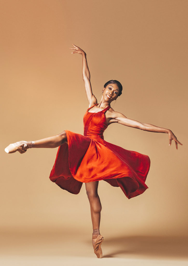 A dancer in a red dress smiles at the camera while in a right leg front attitude on a left diagonal with her arms in an L shape.