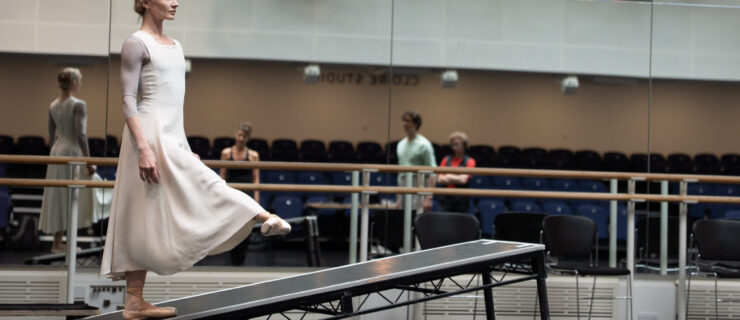 A dancer wearing pointe shoes and a long white dress faces the right side with her arms at her side and right leg extended to the front with a flexed foot. Her standing leg balances on the end of a long skinny black table that sits on a diagonal with the end of it's left two legs cut off. Behind her is a line of black chairs and a ballet bar in front of a full length mirror reflecting a blackbox theater and three other dancers watching her in rehearsal.