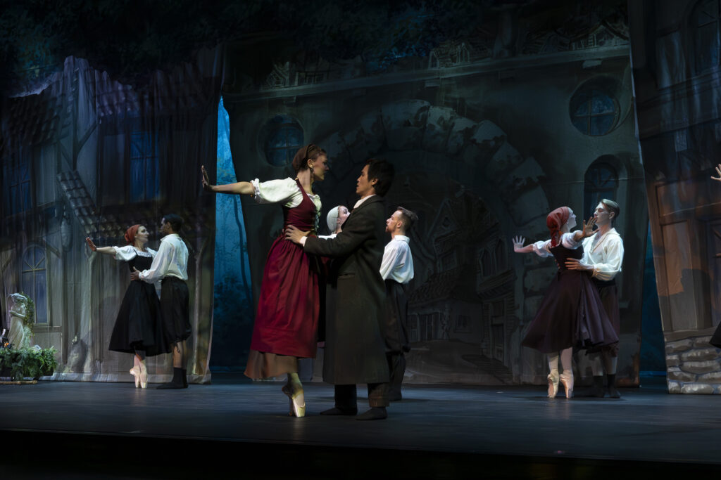 Four couples partner onstage posing in the same position with the male dancers holding the female dancers around their waist, while they balance on releve fifth position with their arms outstretched pushing backwards with flexed hands. The front couple shows the female wearing a red dress with a white shirt underneath while the male dancer wears a long black coat.
