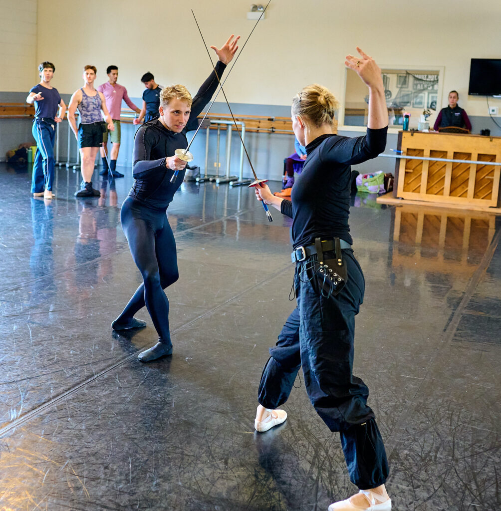 Two dancers in black pose in fourth position with swords crossed during rehearsal.