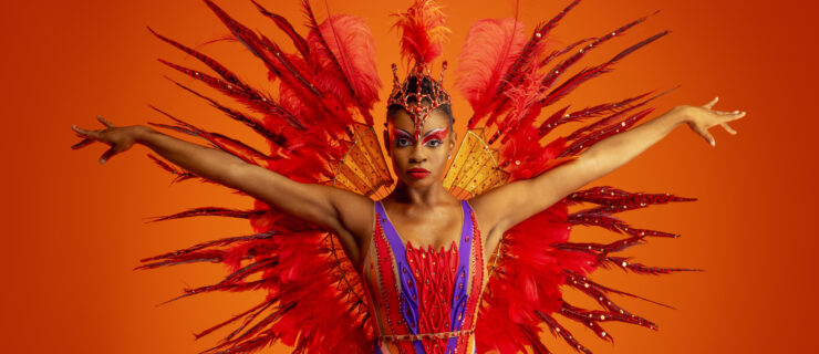 Alexandra Hutchinson is shown from the thighs up, reaching her arms out energetically to the side and looking directly at the camera. She poses in front of an orange backdrop and wears an elaborate costume that includes a red and purple bodice, a jeweled tiara with feather plume, and a set of large, red-feathered wings attached to her back.