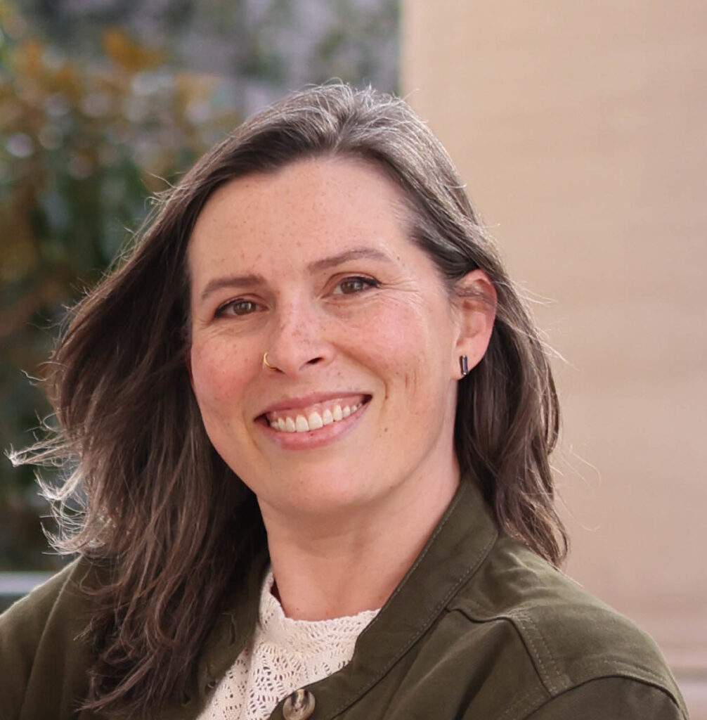 In a headshot, Dr. Jessi White towards the camera with her shoulder-length grey hair down while wearing a white high-neck blouse and green khaki button-up jacket.