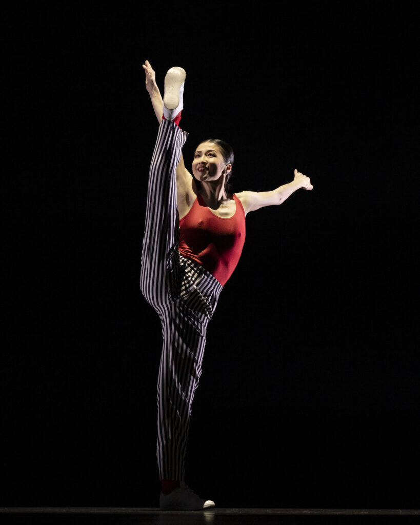 A dancer in a red leotard, black and grey striped pants, red socks, and white shoes poses in mid-battement side with her left leg and left arm straight up and her right arm extended side.
