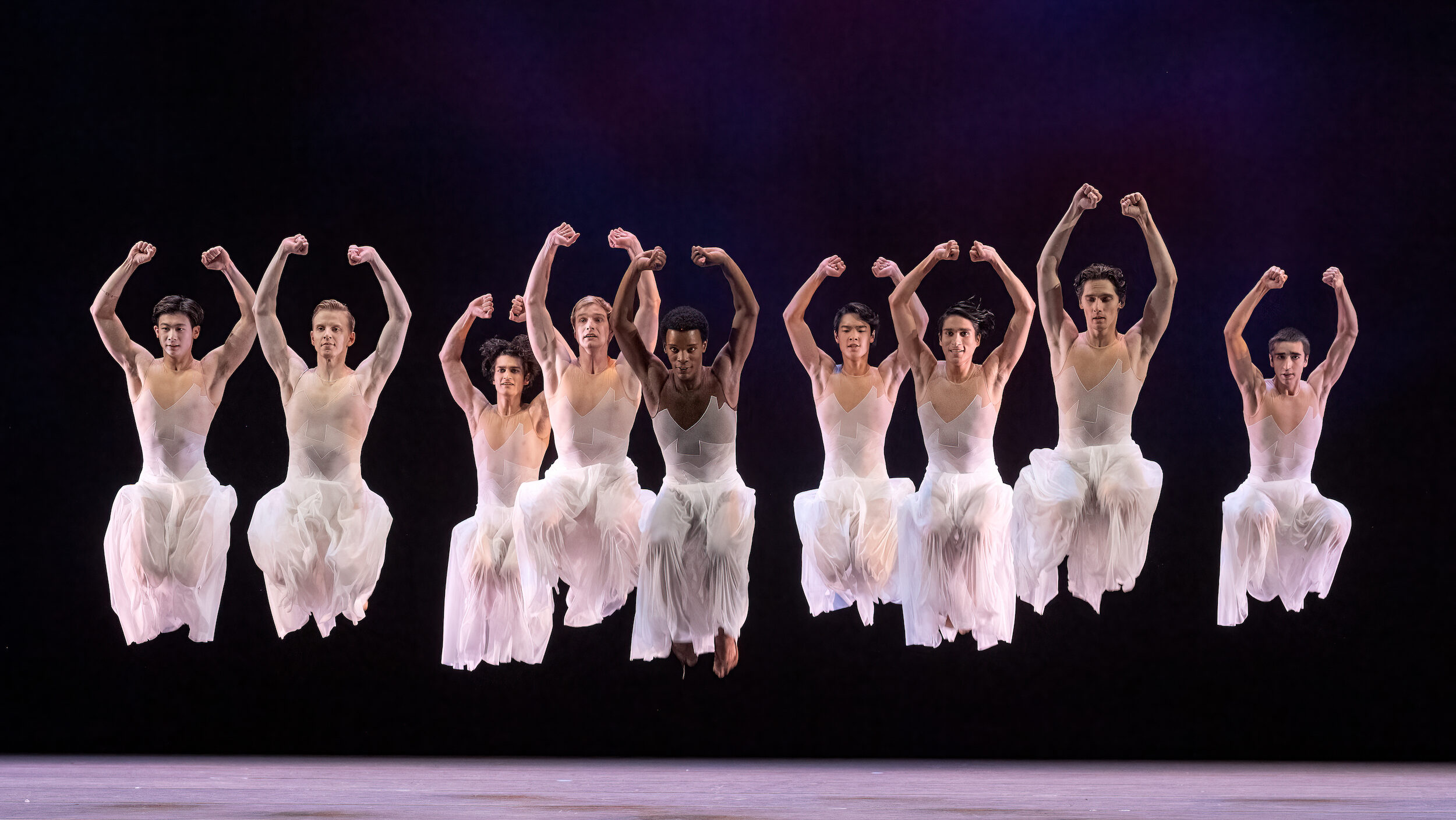 Nine male dancers wearing while sheer flowy pants are suspended in the air mid-jump with legs bend at the knee and arms held above their heads with clenched fists