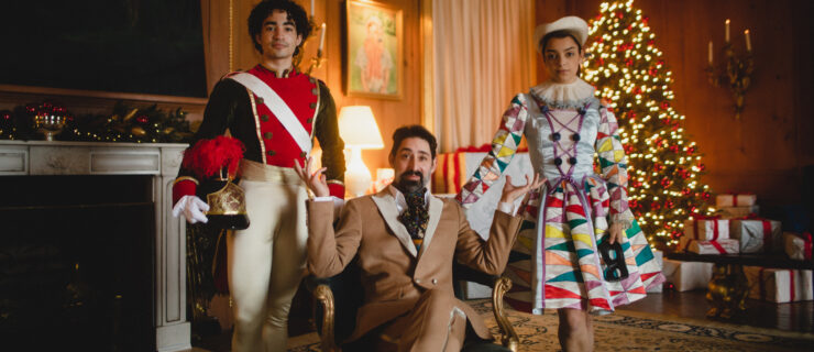In a grand parlor room, a man in a 19th century brown suit sits cross-legged on an antique chair with blue velvet cushions. To his right is a male dancer in a toy soldier costume with white tights, holding his feather-plumed hat in his right hand. To the man's left is a female dancer in a multi-colored Harlequin doll costume. They look directly at the camera with small smiles. In the background is a fireplace and lit up Christmas tree.