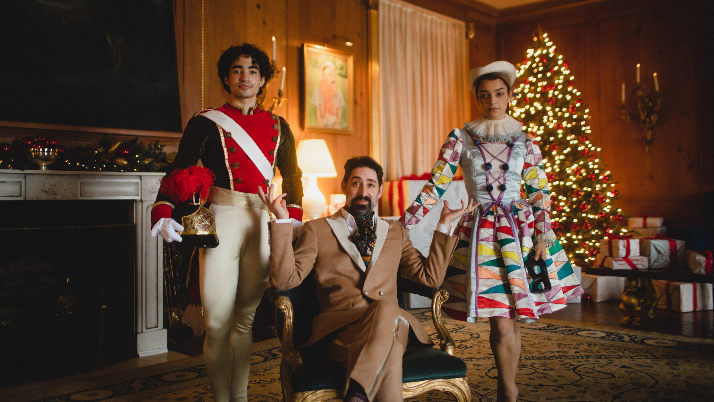 In a grand parlor room, a man in a 19th century brown suit sits cross-legged on an antique chair with blue velvet cushions. To his right is a male dancer in a toy soldier costume with white tights, holding his feather-plumed hat in his right hand. To the man's left is a female dancer in a multi-colored Harlequin doll costume. They look directly at the camera with small smiles. In the background is a fireplace and lit up Christmas tree.