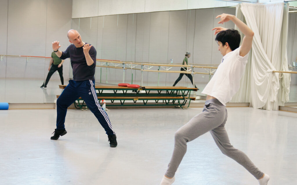 A dancer in grey joggers and a white t-shirt poses in fourth position lunge on releve with arms above his head bent at the elbow. Across from him, Erst Meisner mimics his pose while coaching him in a grey long sleeve shirt and navy adidas pants.