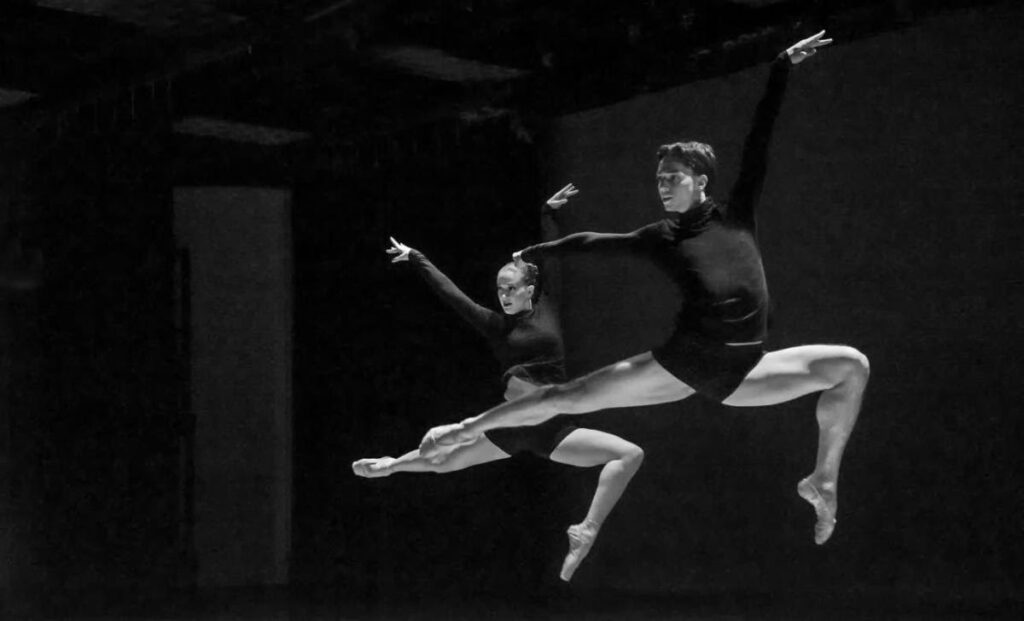 two dancers in black bike shorts and turtlenecks pose in the air mid leap to the right with right legs bent at the knee and left leg extended behind them. Their arms are extended above their heads in a V shape.