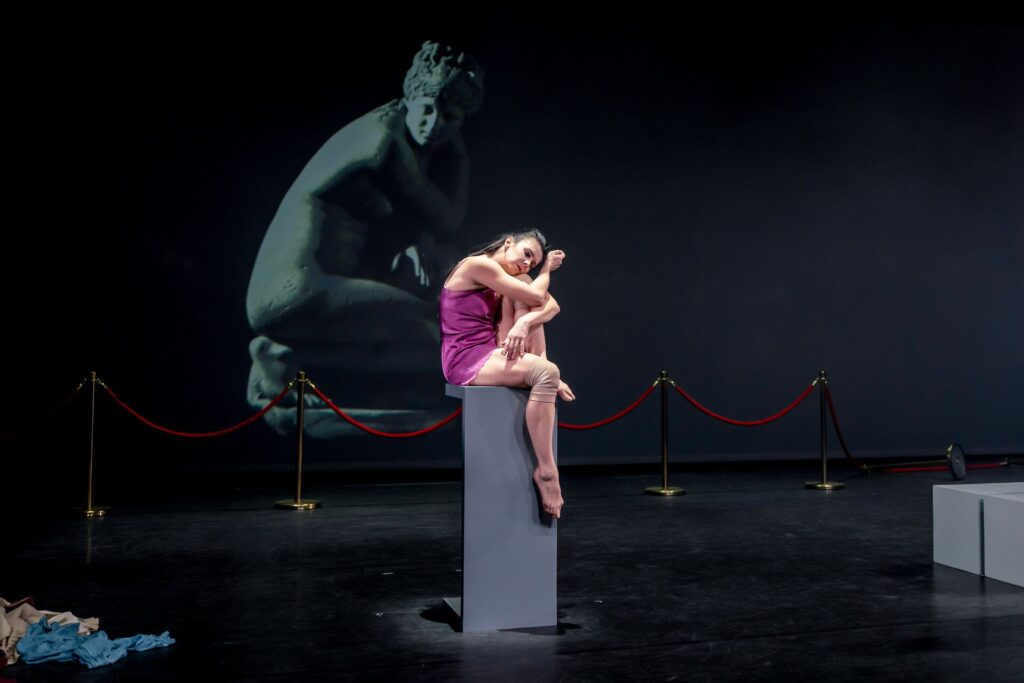 A barefoot dancer in a purple slit dress and knee pads sits on a grey pedestal with her left foot pointed and hanging off the edge while facing the right diagonal and hugging her left knee. Behind her are five gold posts (with the last post knocked over) connected by red velvet ropes in front of a projection of a famous ancient artifact that is a sculpture of a woman crouching in a similar position to the dancer.