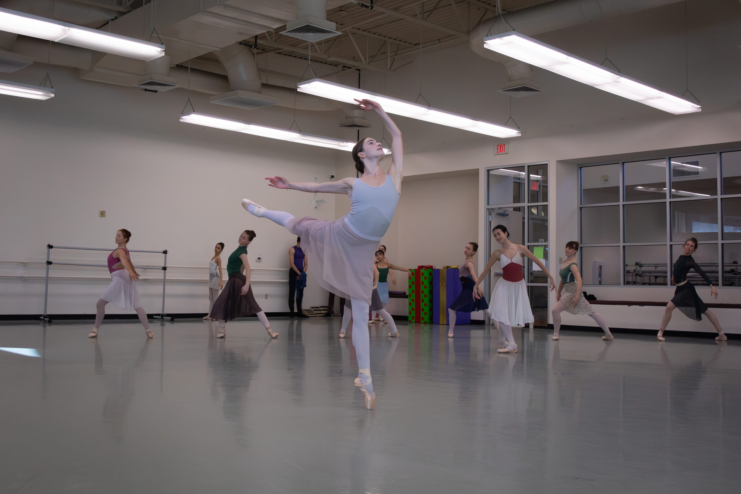 In a ballet studio, a dancer does a piqué arabesque with her arms in high third. She looks up toward her top arm as her rehearsal skirt flows. Behind her, rows of corps dancers pose in a shallow lunge with their arms curved gently behind them.
