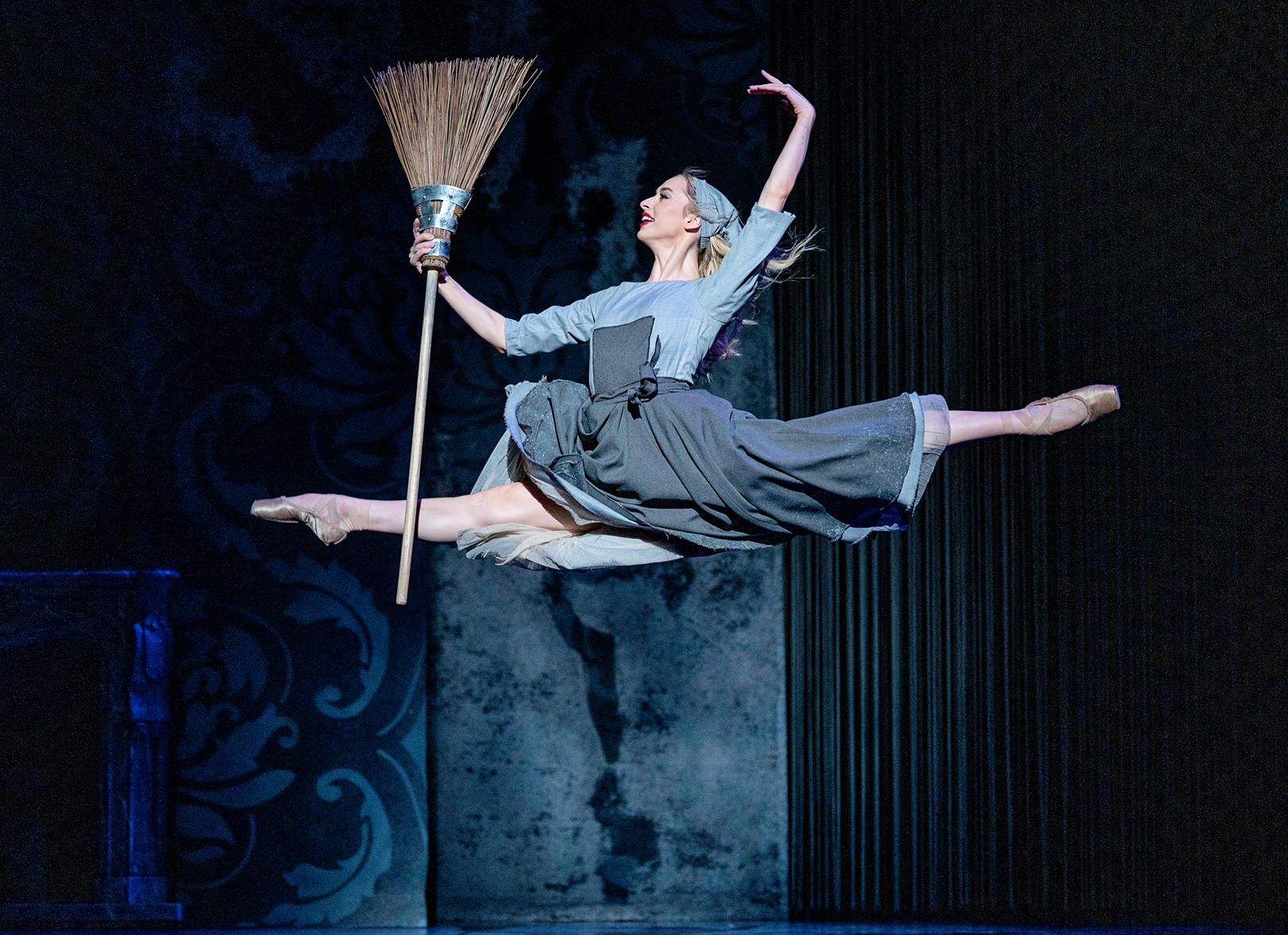 Tomie Lin Kesten, costumed in a plain blue dress and apron, a matching bandana around her head, and pointe shoes, does a saut de chat onstage. She holds a broom in her right hand and looks toward it, and holds her curved left arm up above her head.