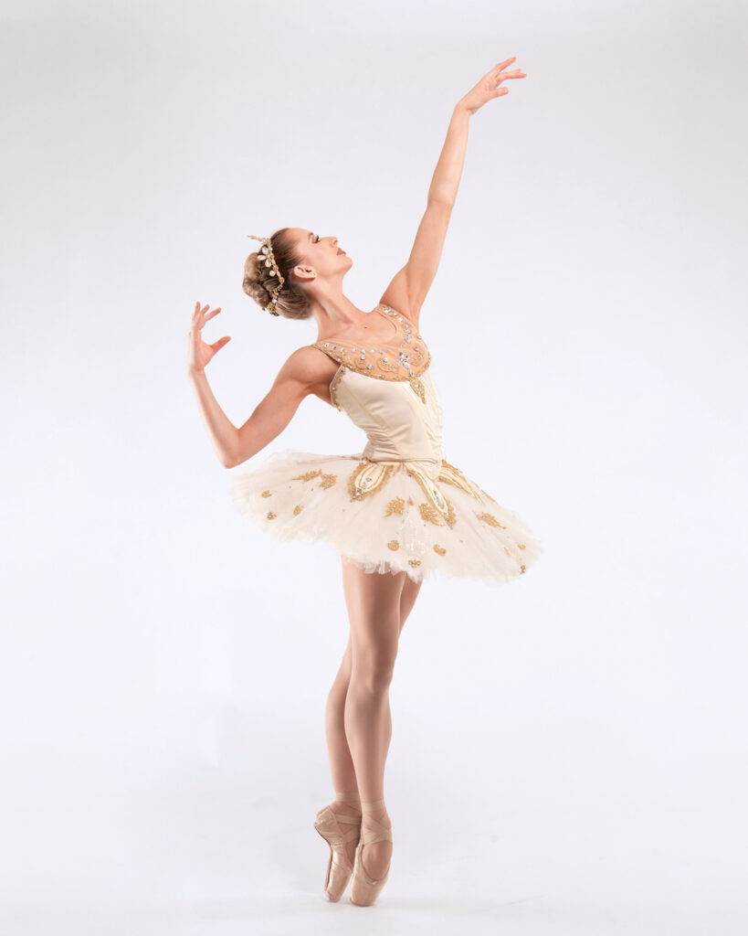Tommie Lin Kesten arches backwards with her right arm extended upwards and her left arm bent at the elbow while in releve fifth position wearing a gold bejeweled tutu.