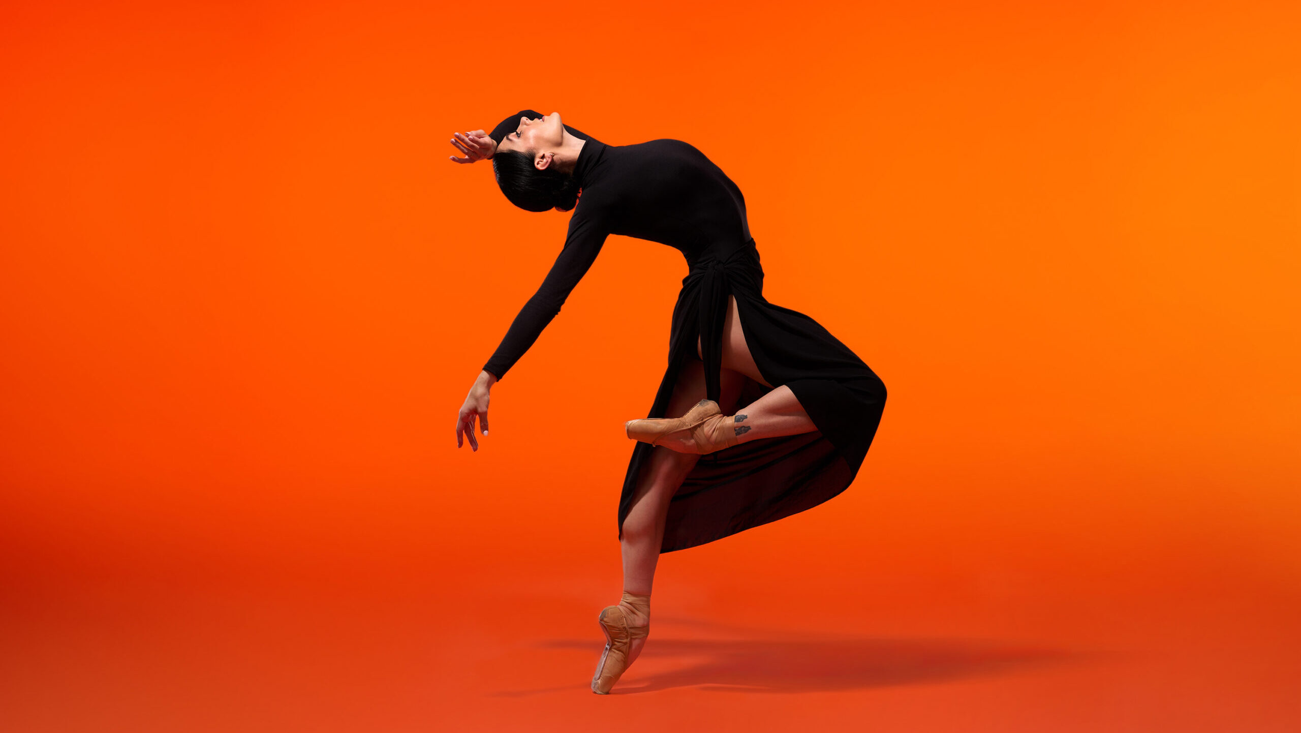 Beckanne Sisk poses en pointe in a turned in passé with her right leg to the right while arching back with her right arm extended towards the floor and her left arm bent to the side, her outer wrist resting on her forehead. She wears nude pointe shoes, a long-sleeve black turtleneck leotard, and a long black skirt tied on her right hip with an open slit against a bright orange backdrop.