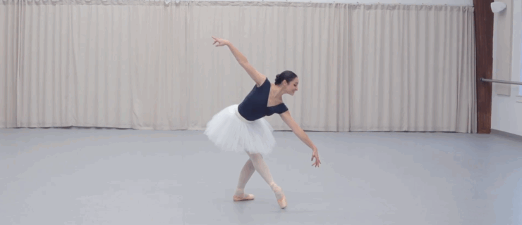 Malena Ani , wearing a cap-sleeved blue leotard, pink tights and pointe shoes, and a white practice tutu, poses with her right leg in tendu croisé devant. She stretches her arms diagonally and looks toward her left hand.