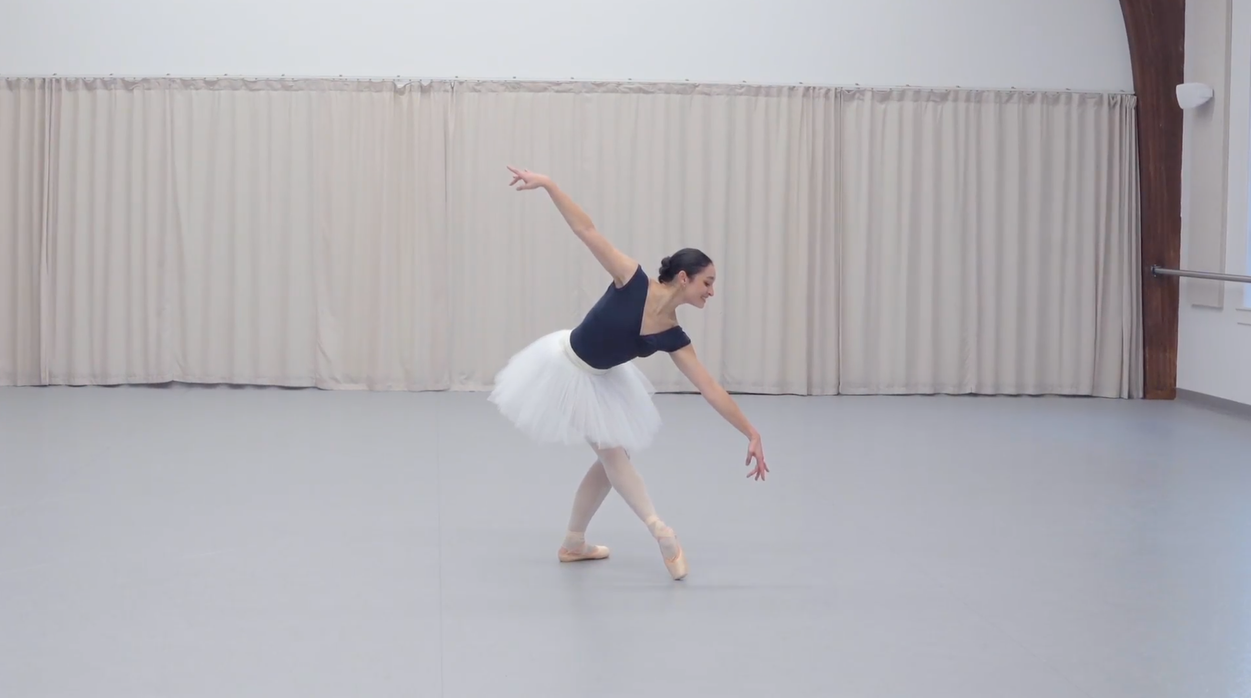Malena Ani , wearing a cap-sleeved blue leotard, pink tights and pointe shoes, and a white practice tutu, poses with her right leg in tendu croisé devant. She stretches her arms diagonally and looks toward her left hand.