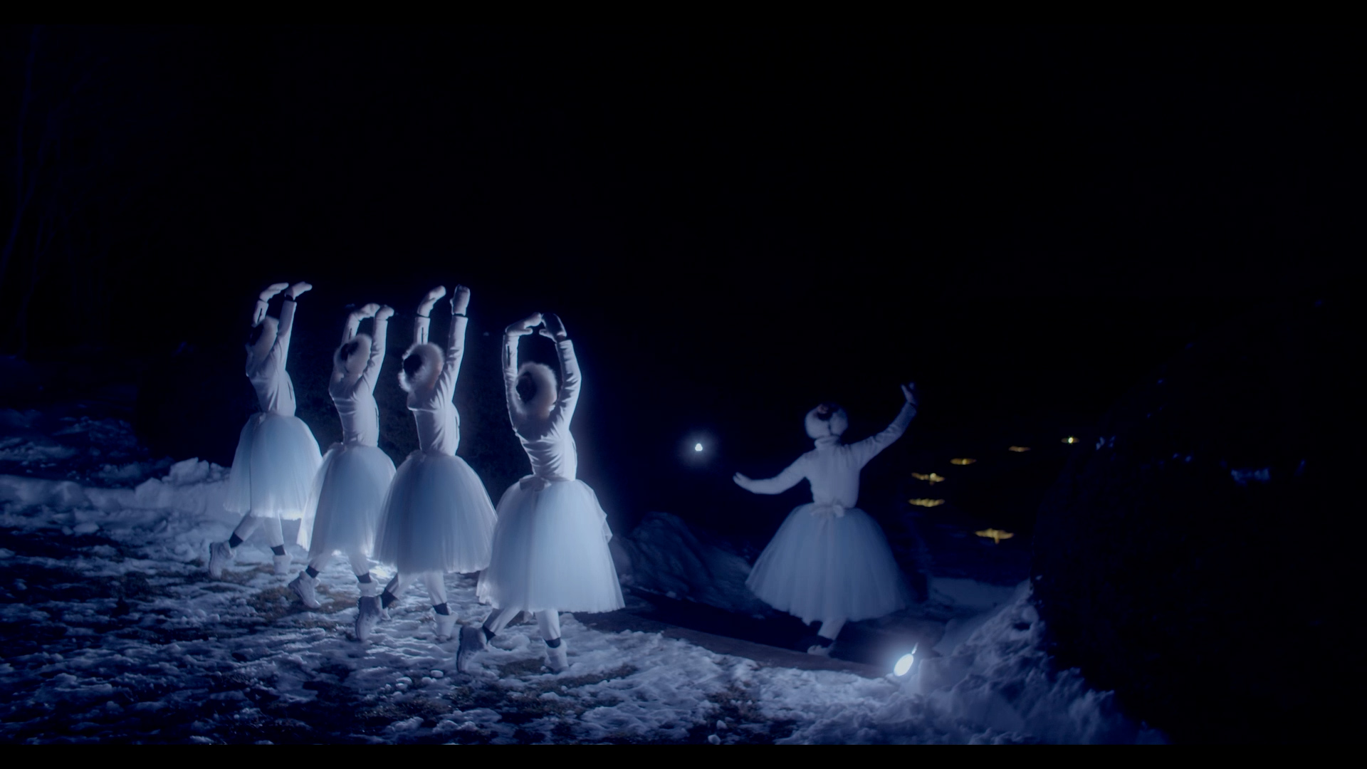 Four ballerinas, shown from behind, pose in b plus with their arms in fifth position en haut while another dances in front of them. They wear white outdoor gear, long white tutus, fuzzy white hats, and white booties, and dance at nighttime in the snow in the glow of ground lights.