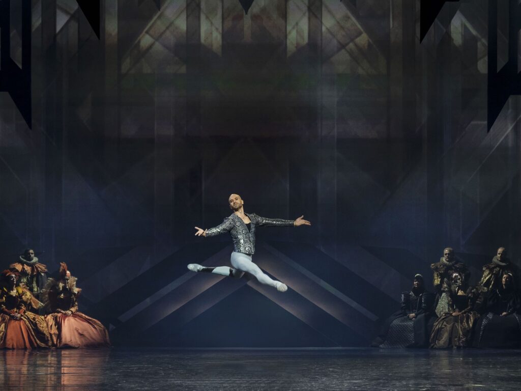 Jonathan Chmelensky wears blue tights and a silver and blue patterned velvet long-sleeved top while suspended mid-leap to the right diagonal with his front right foot extended, his back left foot bent in attitude, and his arms outstretched to the side.
