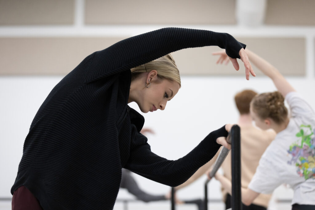 Tommie Lin Kesten wears a black half-zip sweater during company class while arching to the right with her left arm extended over her head towards the barre.