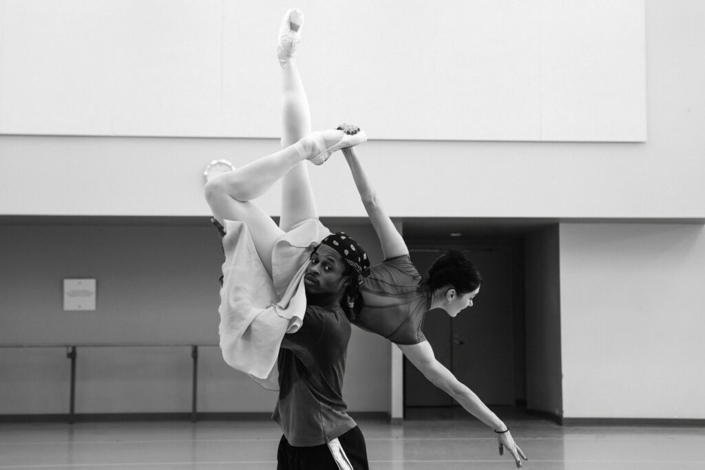 A black and white photo shows Beckanne Sisk faceing away from the camera with in a shoulder lift with Shaakir Muhammad who is looking directly at the camera. She is tilted towards the floor on his left shoulder with her left arm extended downwards to the right and her right arm extended back holding her left foot in passé back while her right leg is straight extended upwards. She wears a long skirt, high neck cap-sleeve leotard, and a french twist in her hair with her skirt hiding Shaakir Muhammad's supporting arms who is pictured, from the pelvis up wearing a long-sleeve shirt and bandana.