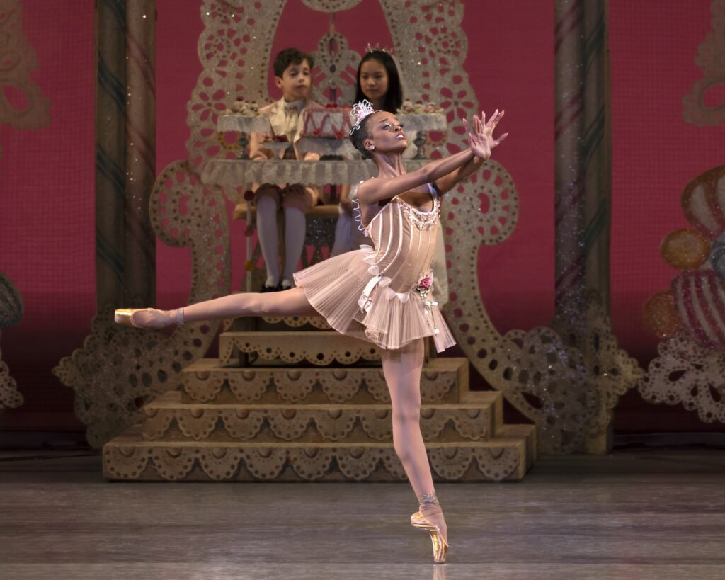 India Bradley, wearing a short, taupe colored tutu, sparkly tiara, brown tights and light brown pointe shoes, does a dynamic piqué arabesque on pointe. She reaches her arms out in front of her and crosses her hands, palms flexed. Behind her, a young boy and girl sit on a filagreed throne behind a small table holding cakes and other sweets.
