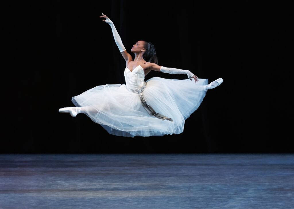 India Bradley, wearing a long white tutu with a taupe sash, long white fingerless gloves, white tights and white pointe shoes, does a saut de chat with her arms in first arabesque. She wears her dark hair in a ponytail, looking up towards her right hand, and dances in front of a black backdrop onstage.
