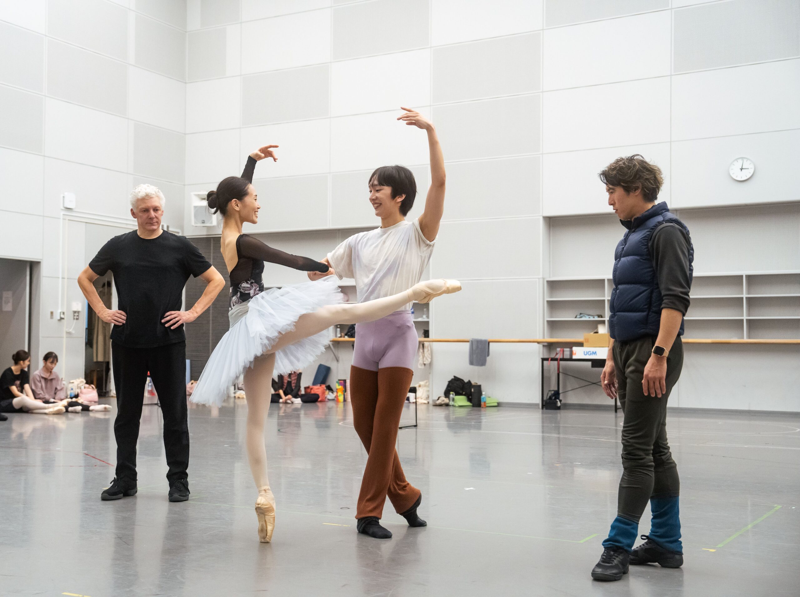 In a large ballet studio, Ayako Ono and Lee Myeonghyun rehearse a pas de deux as Will Tuckett and Yudai Fukouka watch and coach. Ono stands on pointe with one leg lifted devant, holding Myung-hyun's hand as they both lift their other arms to high fifth.