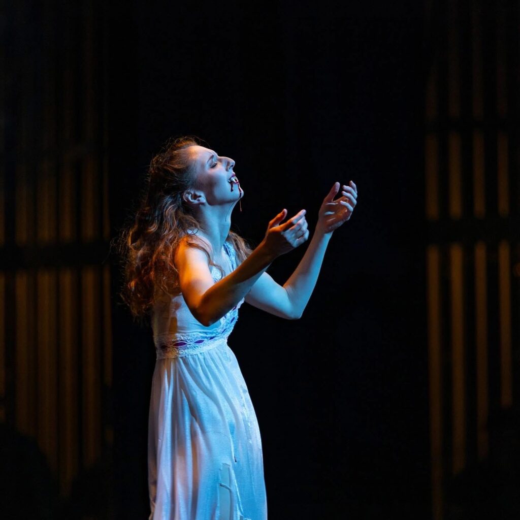 Sarah Kosterman, as Lucy in "Dracula," stands in a white nightgown with blood dripping down her face and neck. She collapses her chest in with anguish, reaching her arms up weakly.