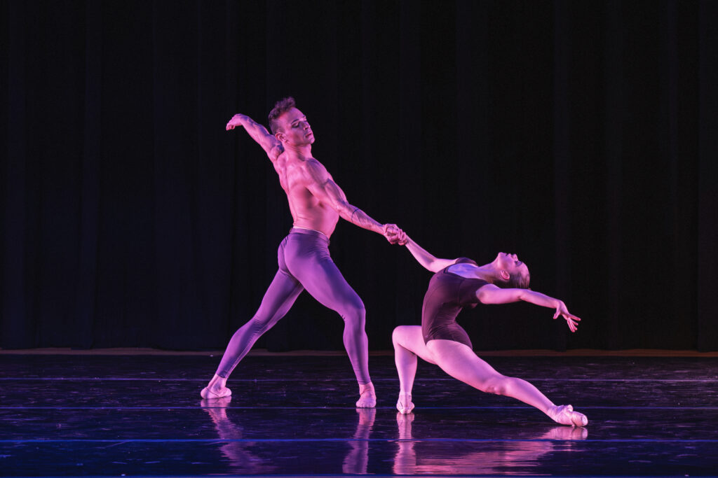 Lorenzo and Perez face each other on a right diagonal, locking right hands while extending their left arms to create a long line. Lorenzo (front, far right) faces towards the back left diagonal while lunging onto her right leg and arching back towards her extended left leg. In opposition, Perez (left and center) faces her on the right diagonal while holding a smaller lunge onto his right front leg. He wears grey tights and Lorenzo wears a dark purple cap-sleeve leotard.