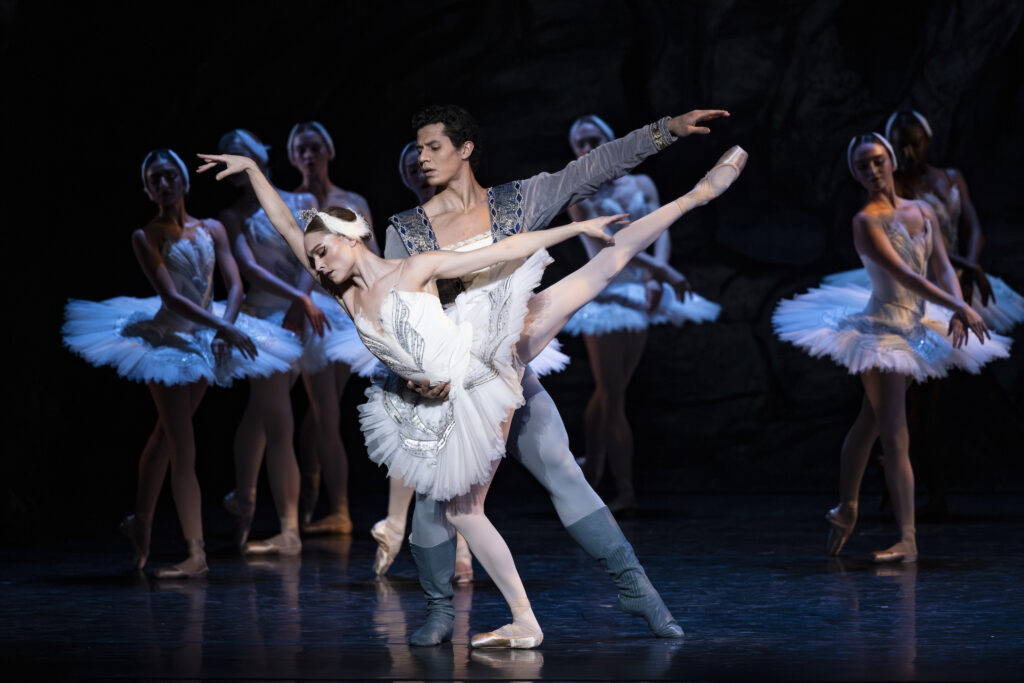 A male and female dancer perform a pas de deux onstage. The ballerina, who wears a white tutu, pink tights and pointe shoes, and a feathered headpiece, does an arabesque allongé towards her right. Her partner, wearing a light blue tunic, tights, and darker blue boots, poses behind her, holding her by her waist with his right hand and extended his let arm out. A corps of dancers in white tutus stand in b plus behind them.