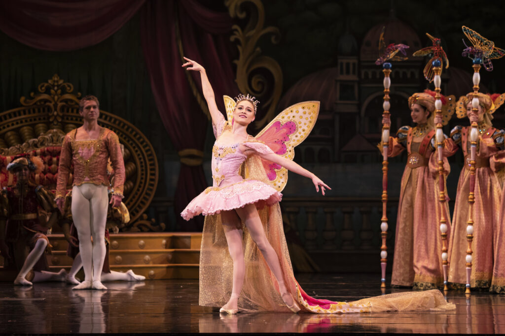 A ballerina stands in tendu derriere effacé with her arms in first arabesque as she looks out towards the audience and smiles. She wears a pink tutu, sparkly tiara, pink tights and pointe shoes, and a gauzy cape with butterfly wings. Other dancers in costu,e stand in the background, watching her.