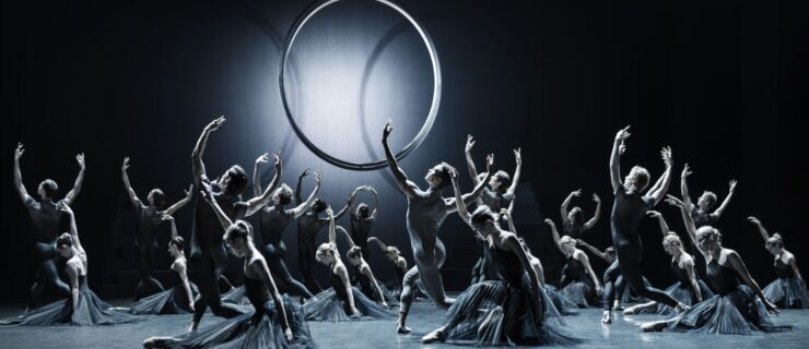 Onstage in stark lighting, a large corps de ballet poses kneeling, sitting, or standing, with many curving their arms upward. Behind them, a large ring is suspended, with a spotlight on it.