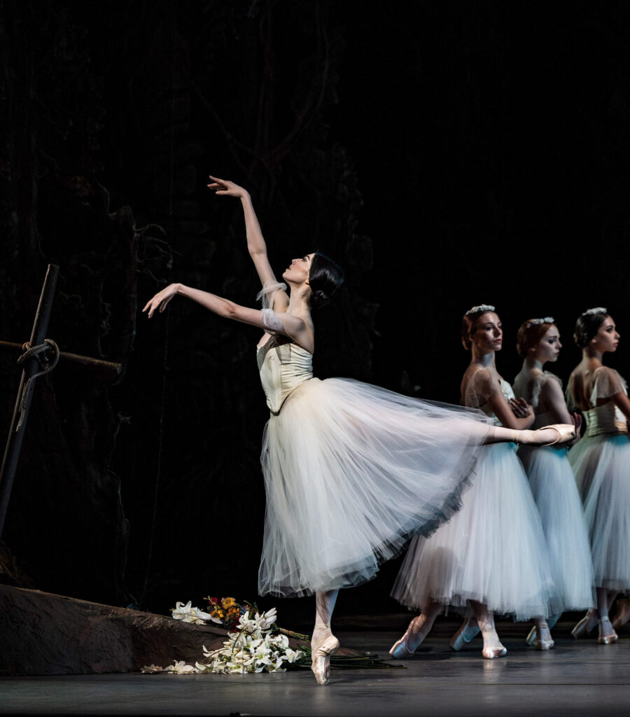 Onstage in front of a dark backdrop with a grave, Chloe Misseldine does a gentle arabesque in profile, arms lifted in front. She wears a long, white Romantic tutu with her hair in a low bun. A line of dancers stand in b-plus on the diagonal behind her.