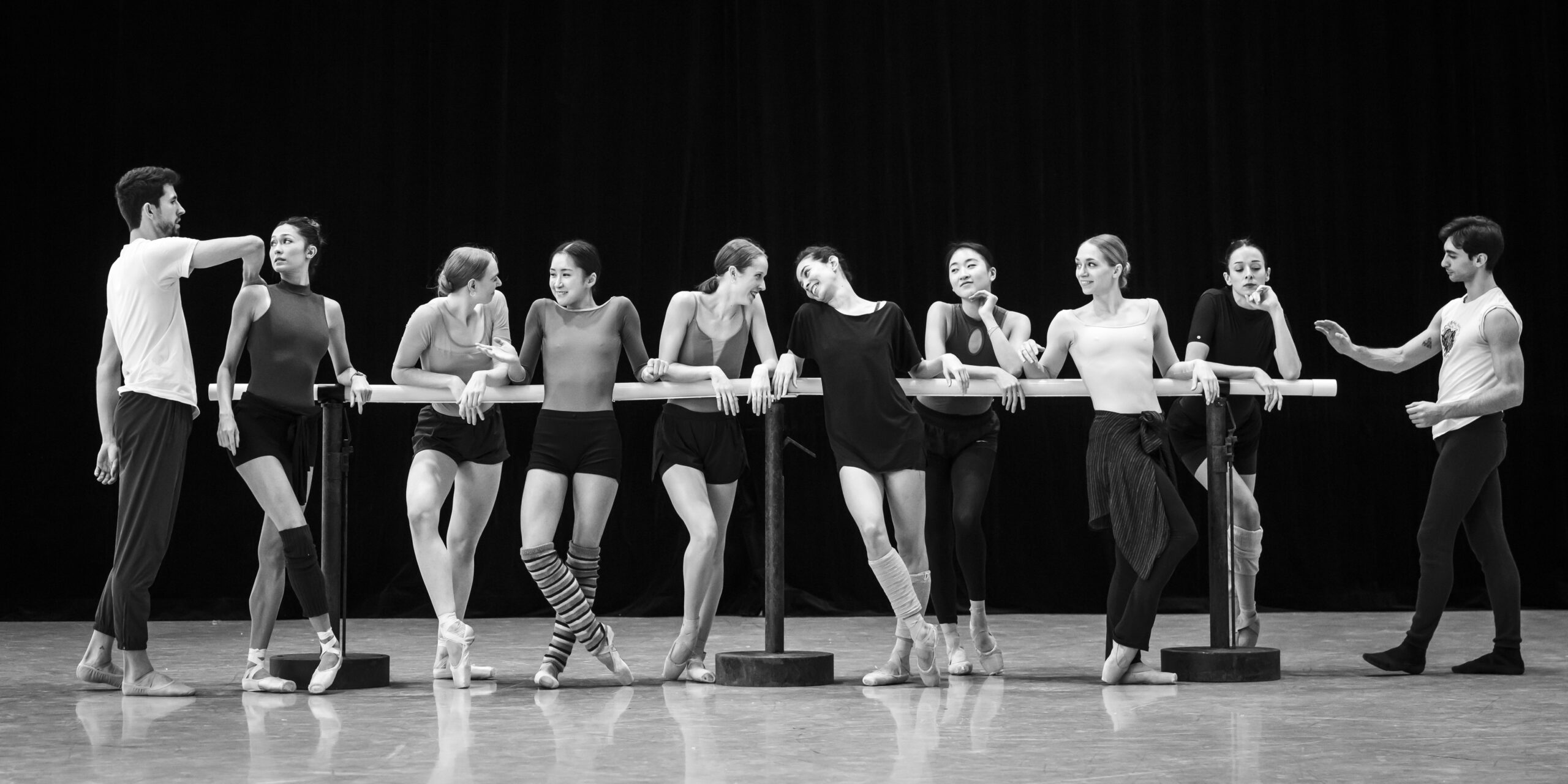 A black and white image of a group of ballet dancers standing together at the barre, casually, speaking and joking.