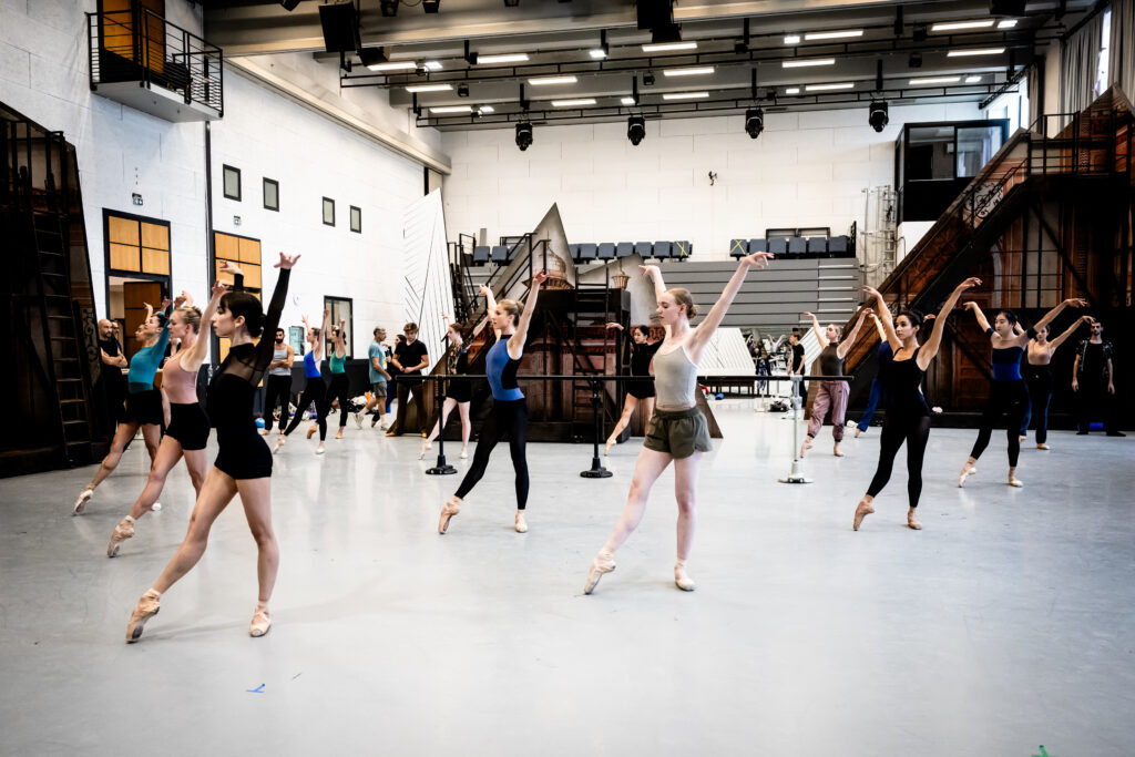 In a large ballet studio, a corps de ballet spreads across the floor in tendu devant, arms up in a "V" shape.