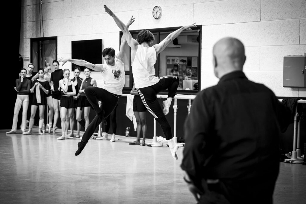 A black and white photo of Jean-Christophe Maillot watching dancers rehearse with his back to the camera. Dancers watch around the studio as two men jump in the retiré position, arms out in a "V" shape.