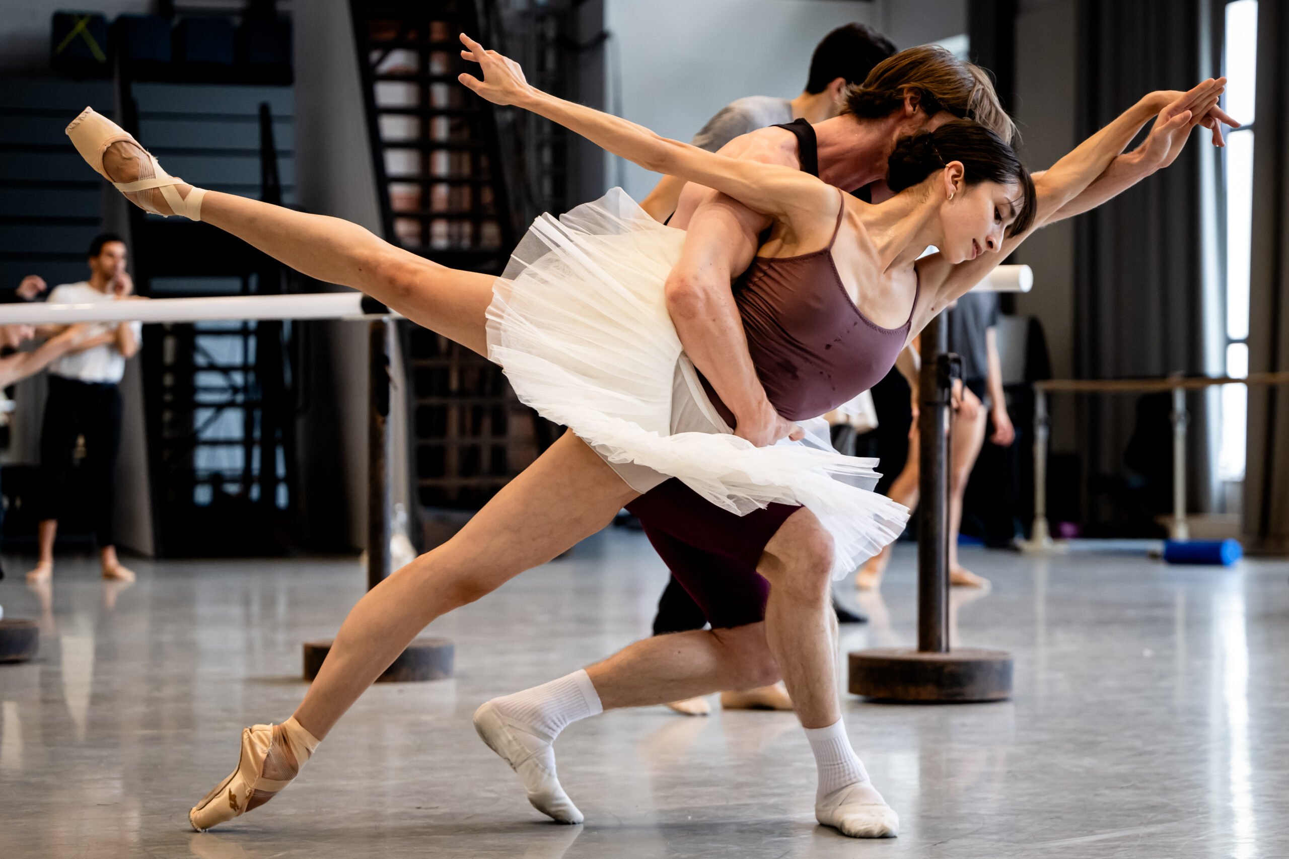 In a ballet studio, two dancers rehearse a pas de deux. The front dancer does a low arabesque as her partner supports her around the waist; her torso faces down as he slides her on the top of her bottom foot's pointe shoe.