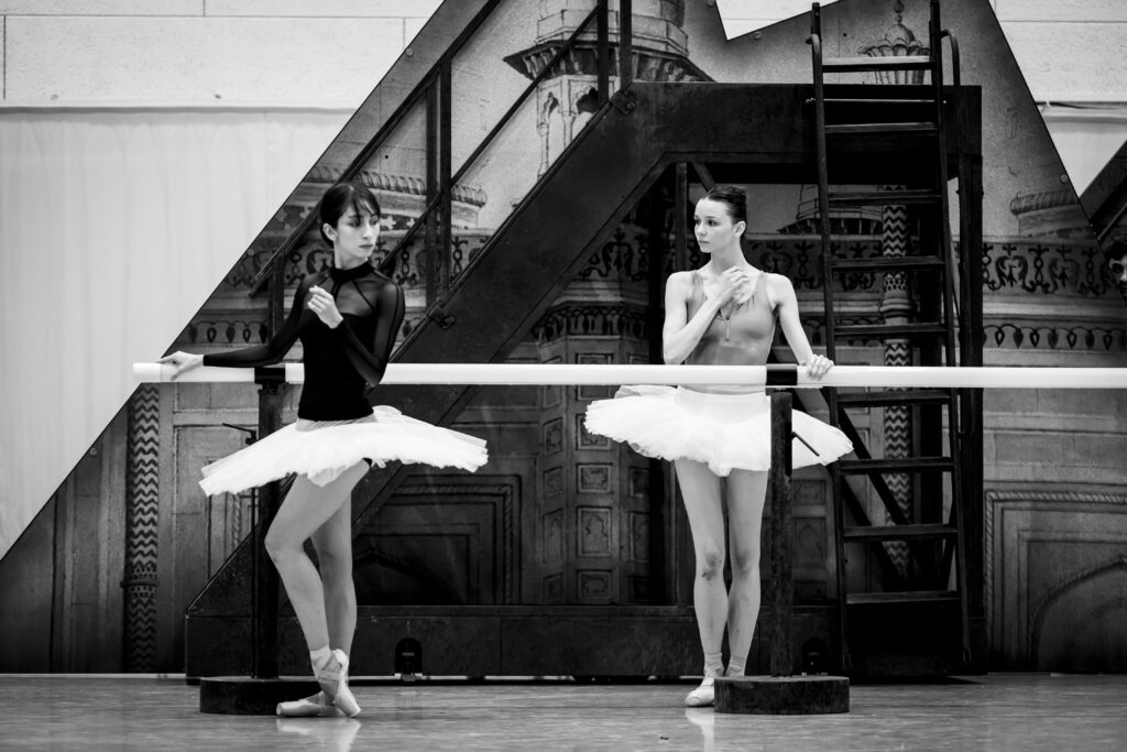 A black and white photo of two ballet dancers in classical tutus standing at the barre, looking at each other warily.