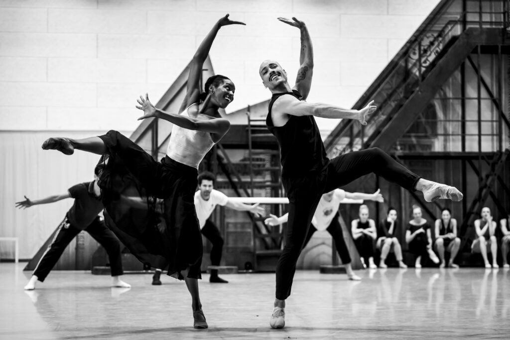 A black and white image of two dancers smiling as they lean toward each other in opposition, one leg held front in attitude and their arms pressing up in a stylized third position. Dancers watch on the floor behind them in the ballet studio, in front of large set pieces.