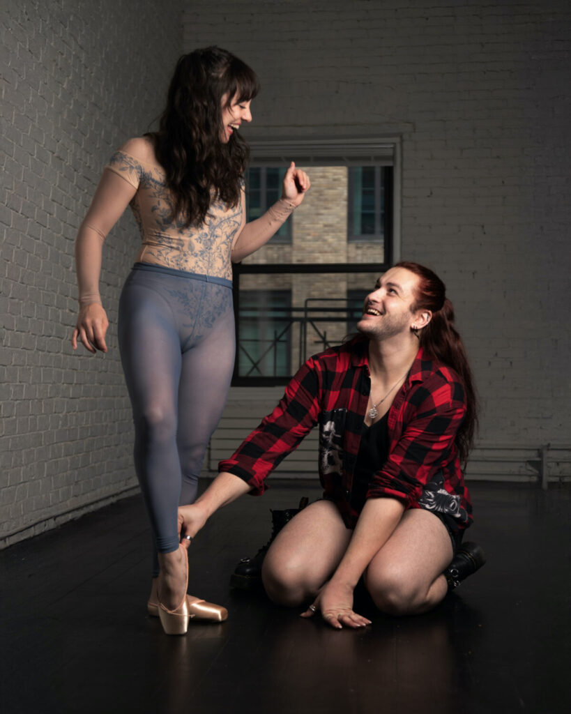 Casey Noble, wearing a red and black flannel shirt and dark shorts, sits on both knees and helps a female dancer as she tries on a new pointe shoe. The woman wears a beige off-the-shoulder leotard with gray lace overlay and gray ankle-length tights.