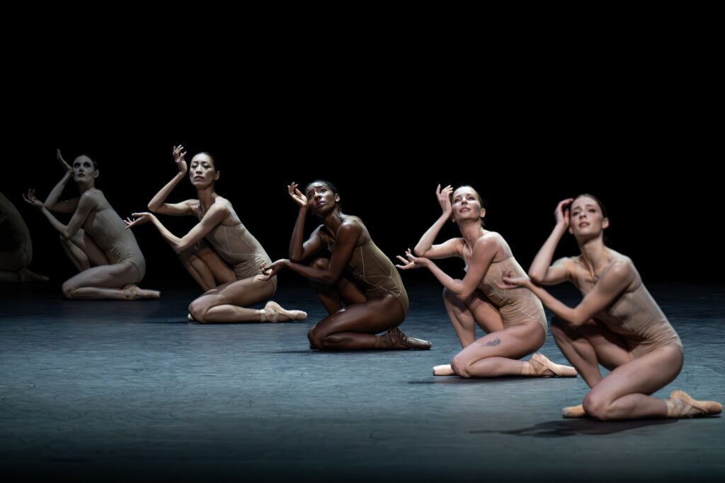 Onstage in darker lighting, five dancers crouch with one leg tucked under, creating a diagonal line across the stage. They tuck their arms in with their bottom hands cradling their top elbows, and their top wrists rest gently against their cheekbones as their fingers flare out delicately. They wear flesh-tone leotards and pointe shoes.