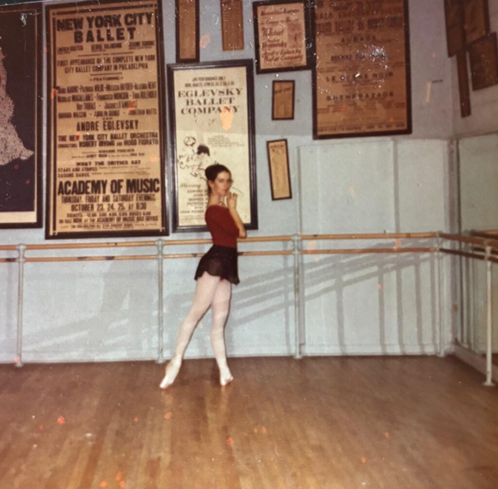 Jennifer Levine poses in profile at the barre in tendu derrierre. She touches her chin with her right hand and looks out toward the camera. She wears a burgundy long-sleeved leotard, black ballet skirt, pink tights and pointe shoes. The studio walls are covered in vintage posters of Eglevsky Ballet and New York City Ballet.