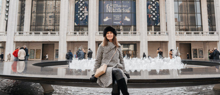 Jennifer Levine, wearing a white and black patterned winter coat, fussy black hat, gray pants and black high-heeled suede boots, and a buff-colored purse slung over one shoulder, sits at the fountain outside of the David H. Koch Theater. She crosses her right leg. A large sign for New York City ballet's "Nutcracker" hangs in the windows of the theater's terrace.